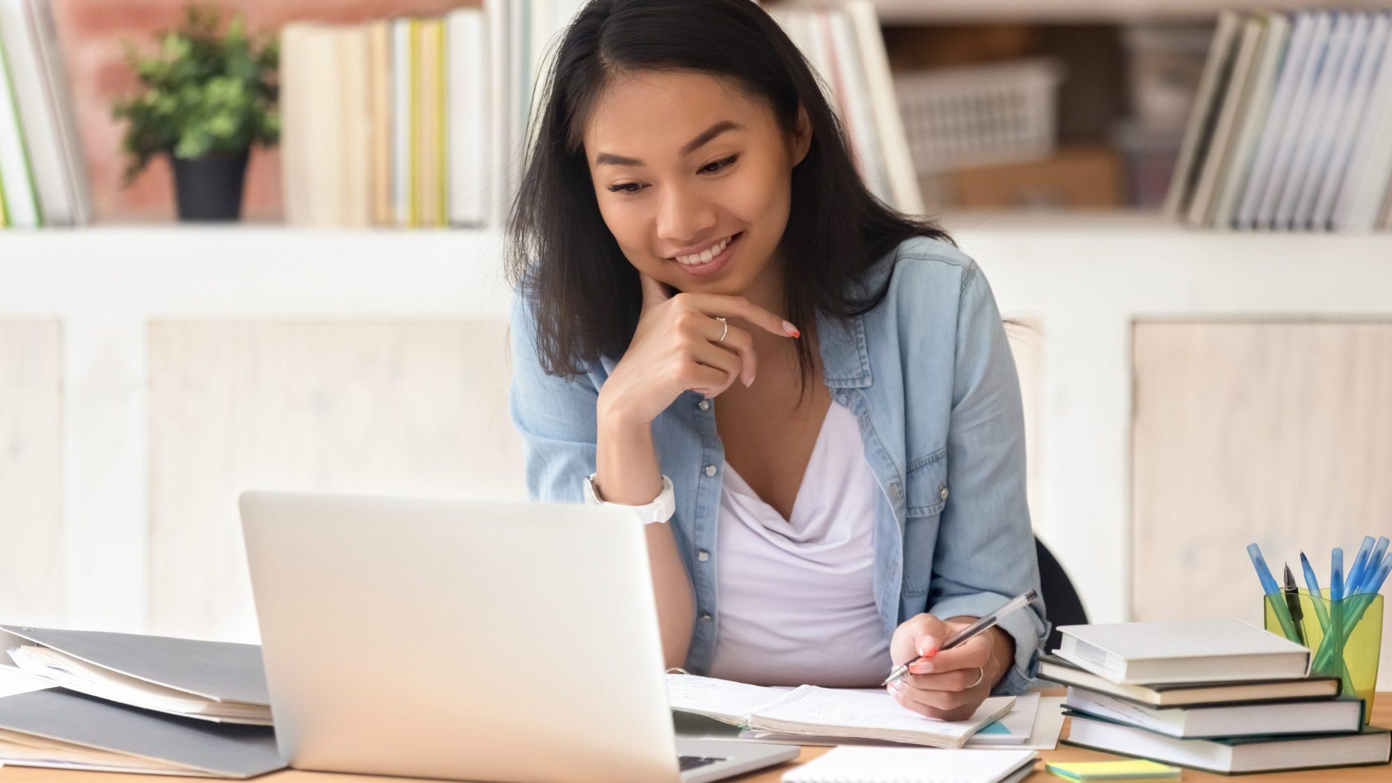 Smiling asian undergraduate teen girl student study in library with laptop books doing online research for coursework, making notes for essay homework assignment, online education e-learning concept